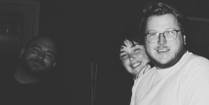 Three people in a dark room, arms around each other.