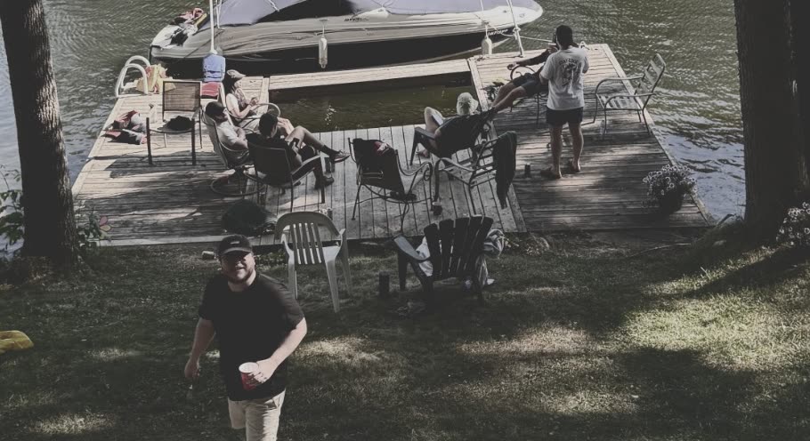 A man in a black shirt walking up the lawn with a drink, six people on the dock behind him.
